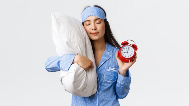 Woman in blue pajamas and sleeping mask, holding pillow and alarm clock with eyes closed